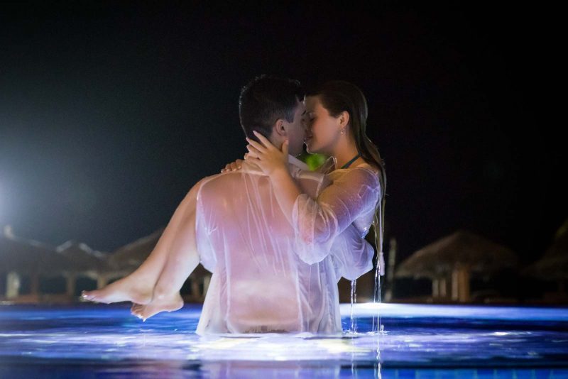 Groom holding up bride in swimming pool at night